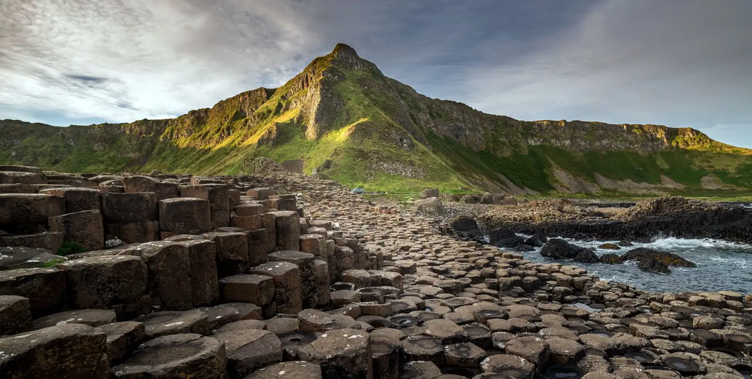 Basalt columns of the Giant's Causeway leading to green cliffs in evening light, on a private Belfast tour